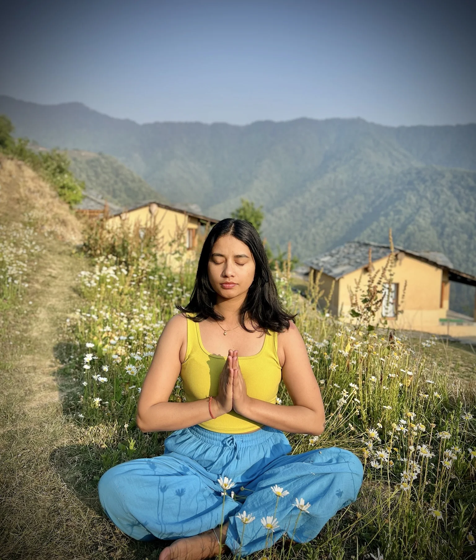 Pooja, founder of Flowherenow, meditating with hands in prayer in a Himalayan meadow